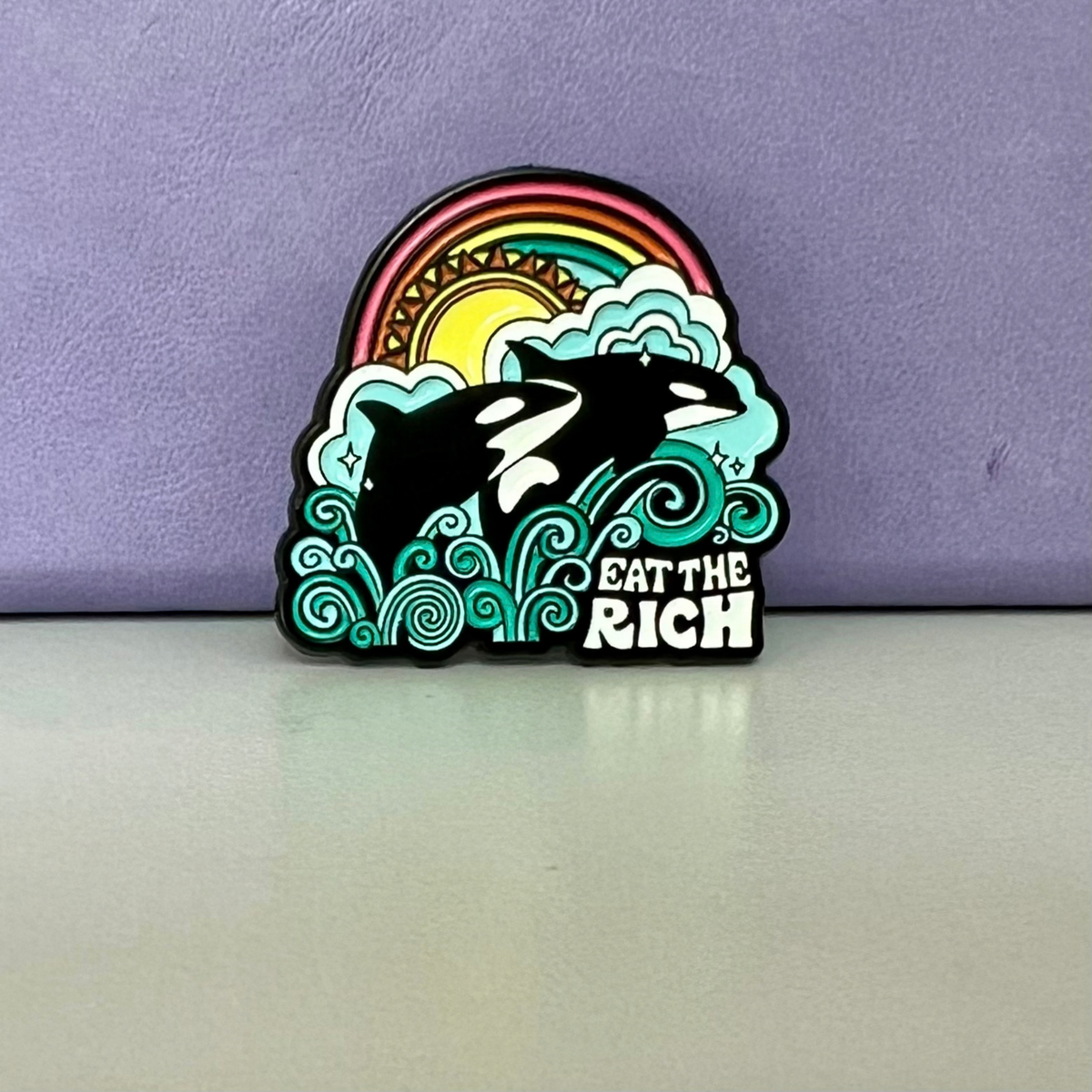 An Eat the Rich Pin featuring two orcas jumping out of stylized waves in front of a rainbow and sun.