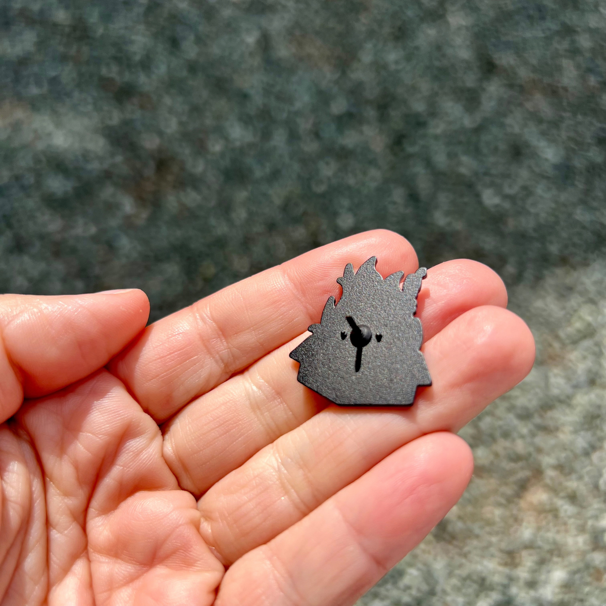 The back of a Fragile But Fearless Pin, a small, flame-shaped black enamel pin with a metal clutch, held in a person’s palm as a statement accessory.