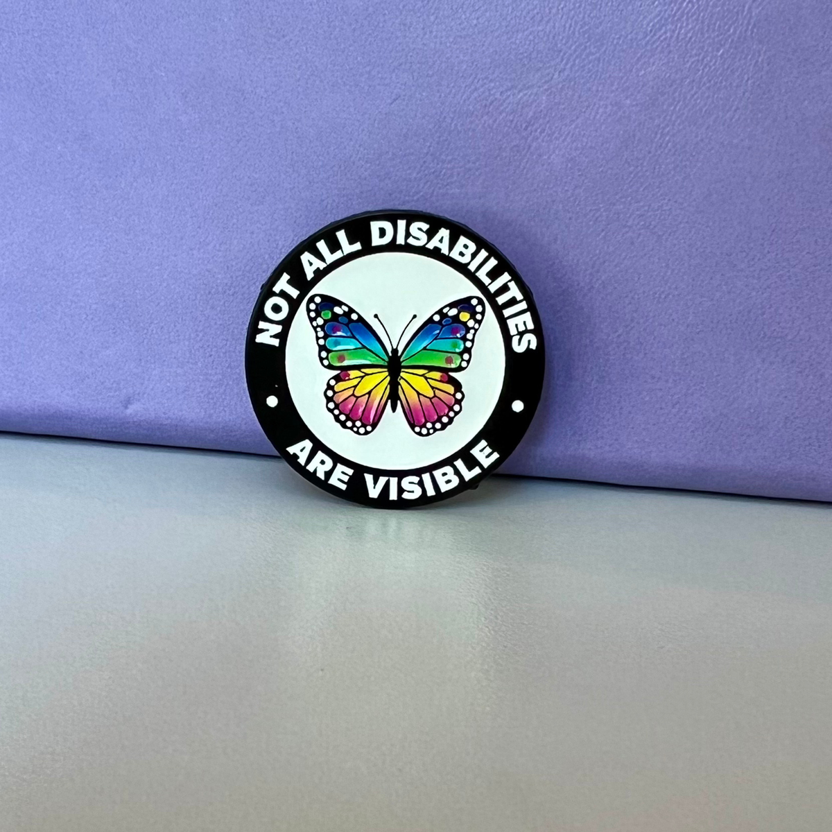 A round enamel pin with a black border and white background, featuring a colorful rainbow butterfly in the center and the text 'NOT ALL DISABILITIES ARE VISIBLE' in white lettering, resting on a light gray surface against a purple background.