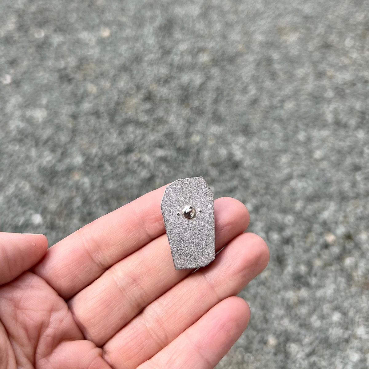 A hand holds the Still Alive! Just Dead Inside Pin by RainBee Designs LLC—a small, metallic coffin-shaped enamel pin with a fastener on the back. This dark humor accessory is displayed against a textured gray surface.