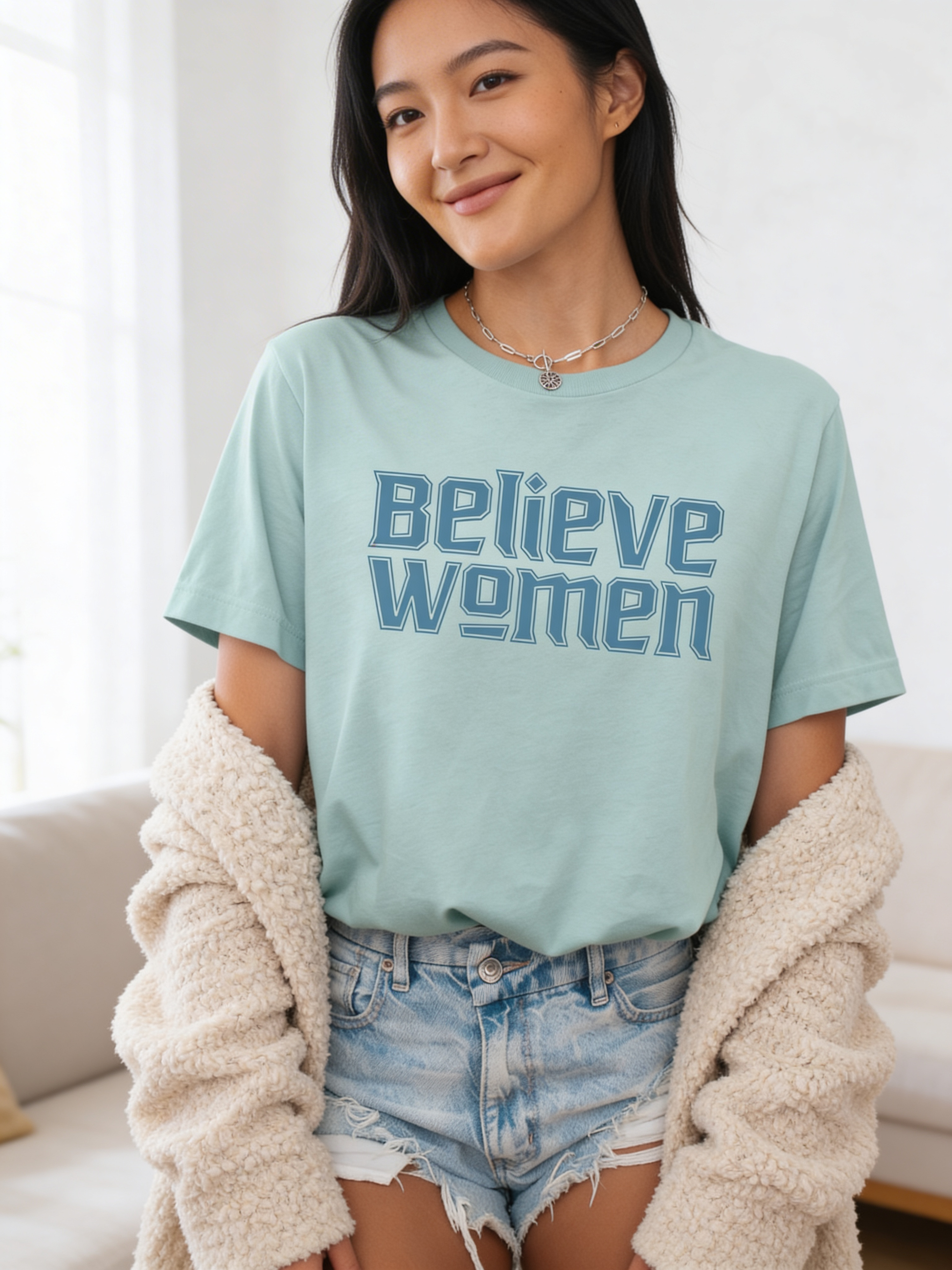 A woman confidently smiles in a dusty blue Believe Women Shirt by RainBee Designs LLC, styled with denim shorts and a cream cardigan draped over her arms.