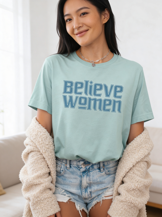 A woman confidently smiles in a dusty blue Believe Women Shirt by RainBee Designs LLC, styled with denim shorts and a cream cardigan draped over her arms.