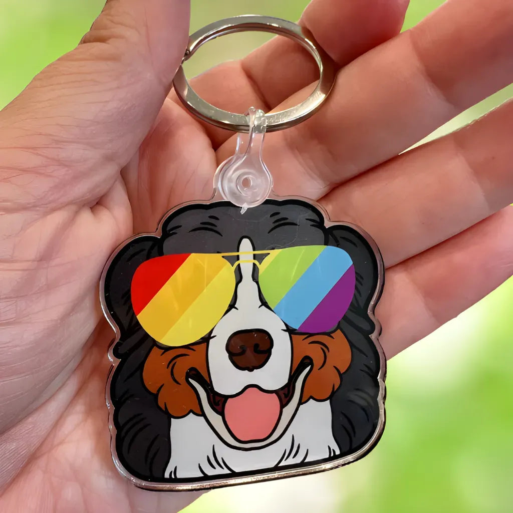 A hand holds the Berner Keychain by RainBee Designs LLC, showing a cartoon Bernese Mountain Dog with rainbow sunglasses and a big smile, set against a blurred green background.