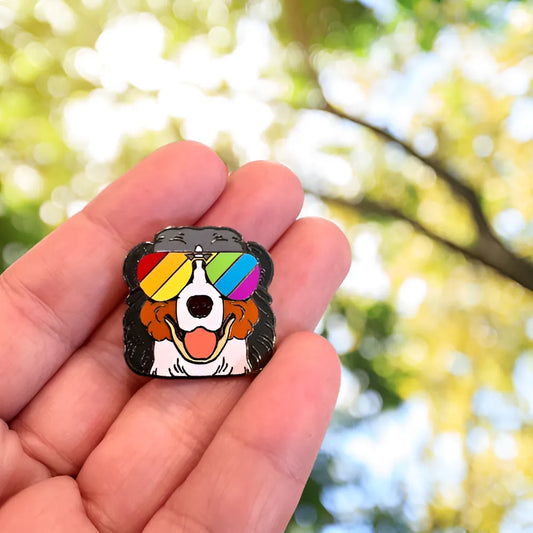 A hand holds the Berner Enamel Pin by RainBee Designs LLC, showing a Bernese Mountain Dog in rainbow sunglasses, with a sunlit background of green and yellow foliage blurred behind.