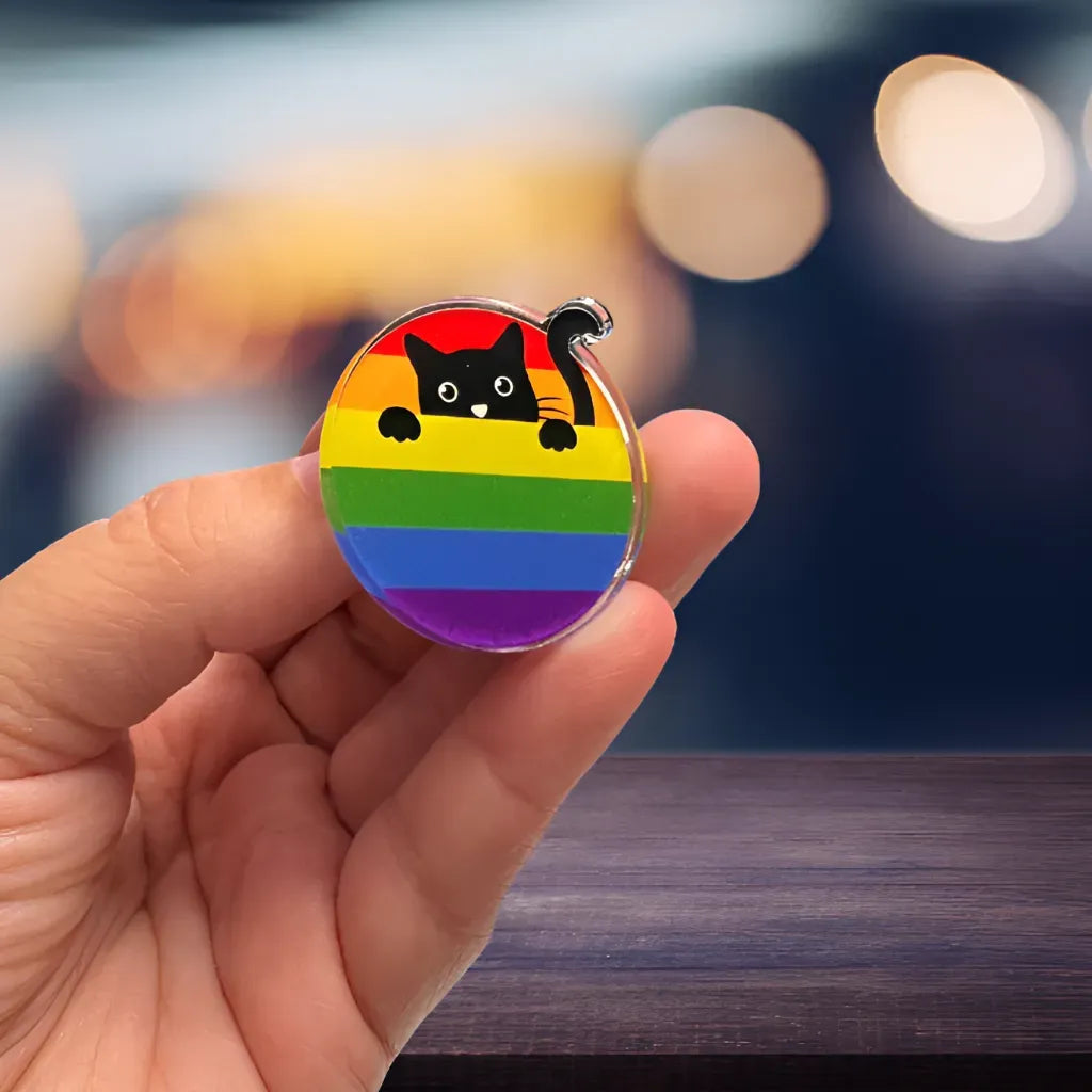 A hand displays the Acrylic Cat Pin by RainBee Designs LLC—a black cat peeking over rainbow stripes, perfect for celebrating LGBTQ+ pride. The softly glowing background is blurred.