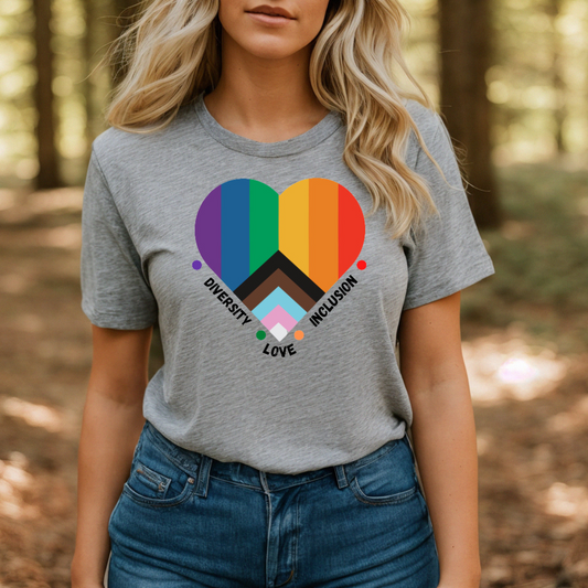 A person stands in a forest wearing the Pride Heart T-Shirt (Grey) by RainBee Designs LLC, which features a heart-shaped progressive pride flag with the words "Diversity, Love, and Inclusion" printed below—a celebration of diversity and love amidst nature.