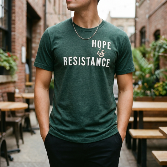 A person stands outdoors in a green Hope is Resistance T-Shirt by RainBee Designs LLC, making a bold statement in style. The premium triblend tee is showcased against a backdrop of wooden tables, plants, and brick buildings for a casual urban vibe. This shirt makes the perfect activist gift for anyone passionate about standing up for change.