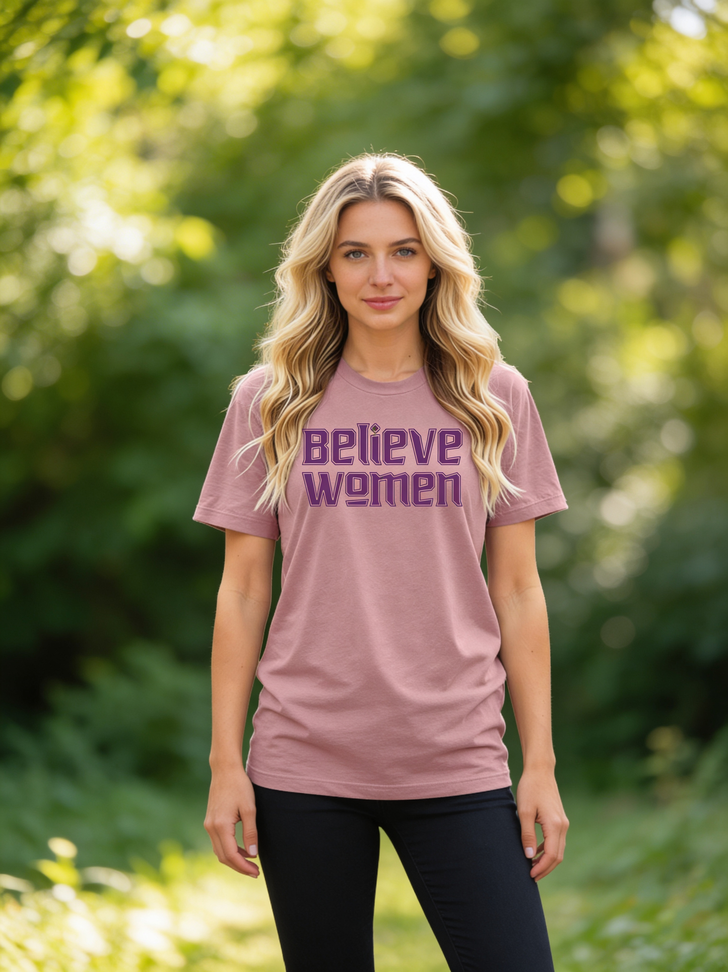 A woman with long blonde hair stands outdoors in sunlit greenery, wearing the Believe Women Shirt by RainBee Designs LLC in orchid—a bold feminist shirt featuring “Believe women” in purple letters. She looks confidently at the camera.
