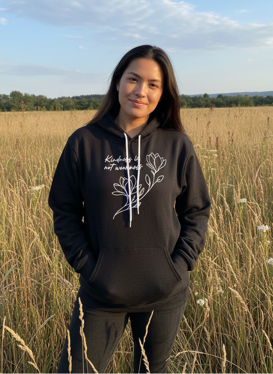 A woman with long dark hair stands in tall grass beneath a blue sky, wearing the "Kindness is Not Weakness" hoodie by RainBee Designs LLC, featuring a white floral design and text on the front, and smiles softly at the camera.