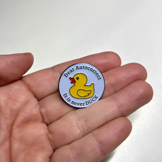 A person’s palm holding a round Dear Autocorrect It Is Never Duck Pin, featuring a yellow rubber duck and the text Dear Autocorrect, it is never DUCK printed around the border.