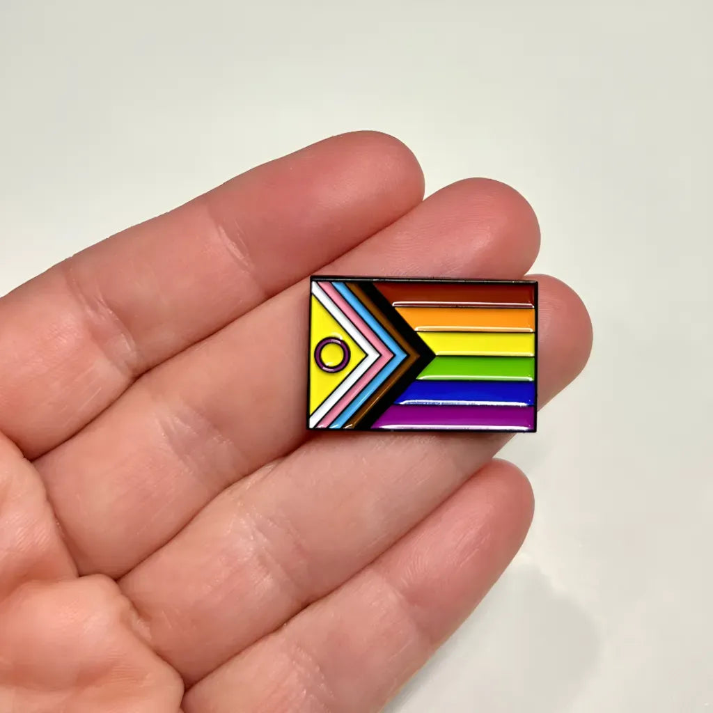 A rectangular Progress Pride Flag (with Intersex) Pin featuring the Progress Pride flag with a yellow circle and purple ring, held in a person’s palm.