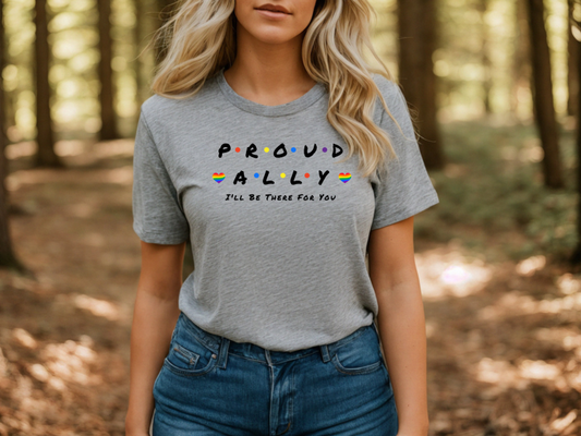 A person stands in a sunlit forest wearing the Proud Ally T-Shirt by RainBee Designs LLC, a gray Friends-inspired shirt featuring colorful text that reads PROUD ALLY and I’LL BE THERE FOR YOU, accented with small rainbow stripes on each side—a perfect allyship apparel choice for showing your support.