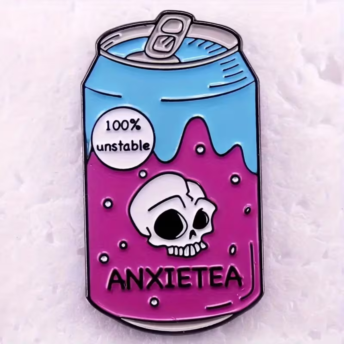 A enamel pin shaped like a soda can labeled 'ANXIETEA' with a blue top half, pink bottom half, a white skull graphic, and a white circle reading '100% unstable'.