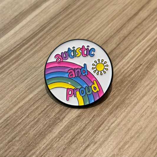 The Autistic and Proud Pin by RainBee Designs LLC is a round neurodiversity enamel pin featuring a rainbow, sun, and colorful “autistic and proud” text, shown lying on a light wood surface.