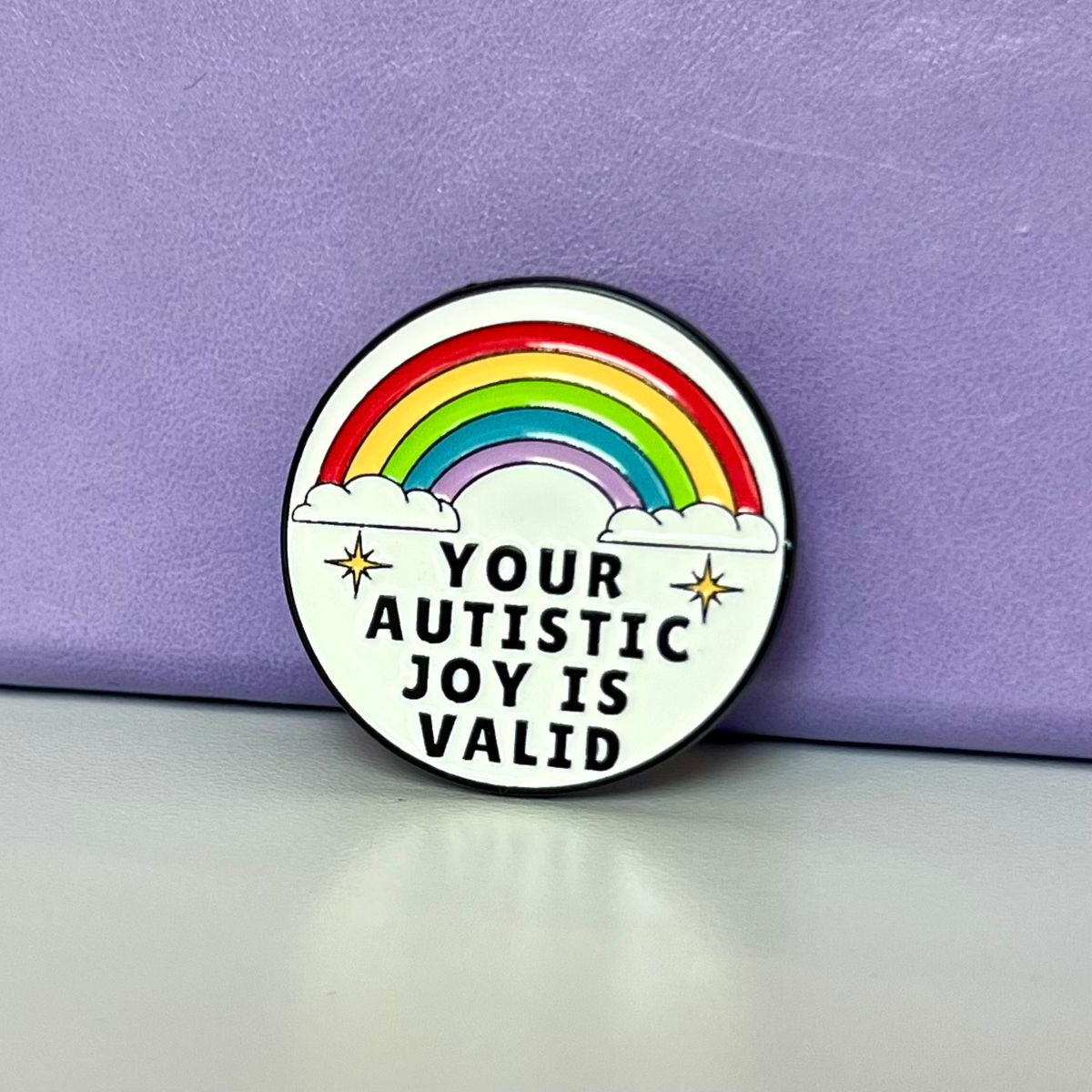 A circular enamel pin featuring a rainbow, clouds, and the text 'YOUR AUTISTIC JOY IS VALID' on a white background, leaning against a purple textured backdrop.