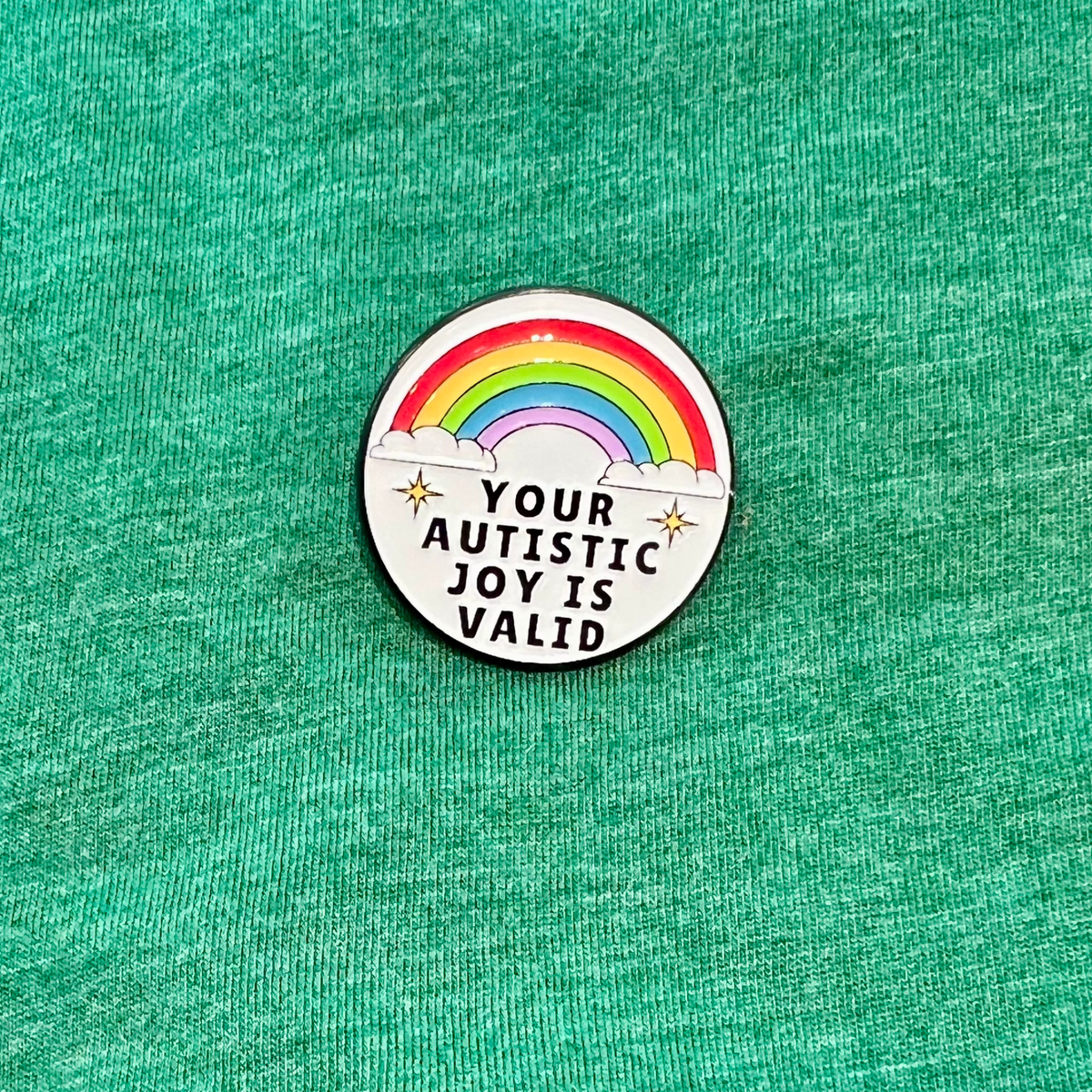 A circular enamel pin featuring a rainbow and the text 'YOUR AUTISTIC JOY IS VALID' with small stars, placed on a textured green fabric background.