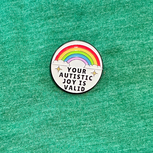 A circular enamel pin featuring a rainbow and the text 'YOUR AUTISTIC JOY IS VALID' with small stars, placed on a textured green fabric background.
