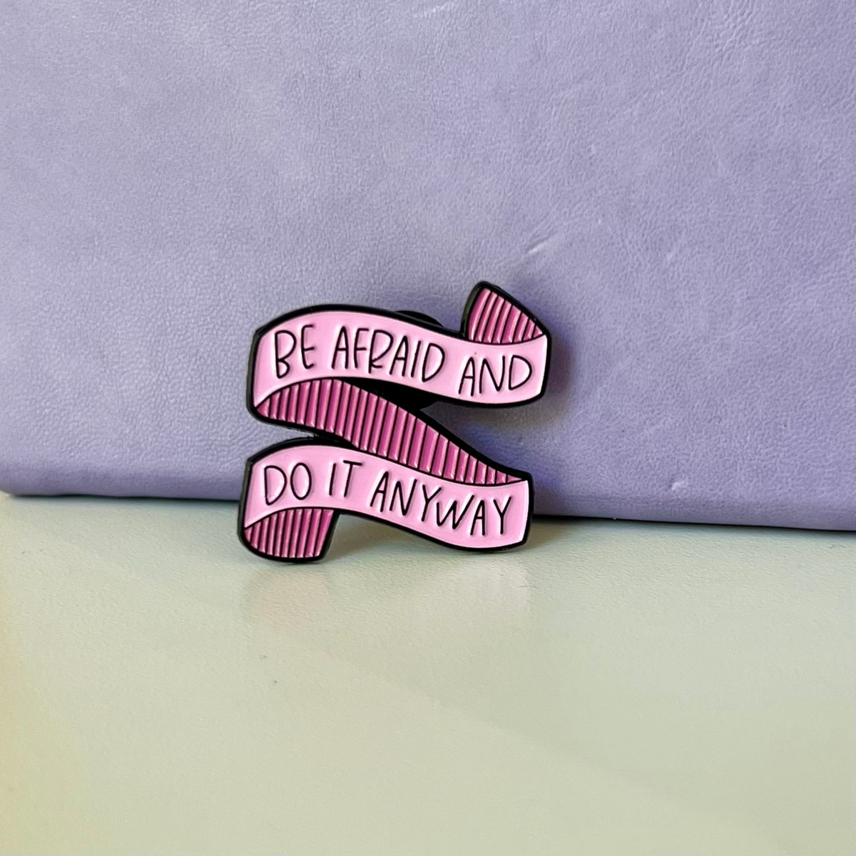 A pink enamel pin shaped like a ribbon with the text 'BE AFRAID AND DO IT ANYWAY' in black outline lettering, displayed on a light grey surface against a lavender background.