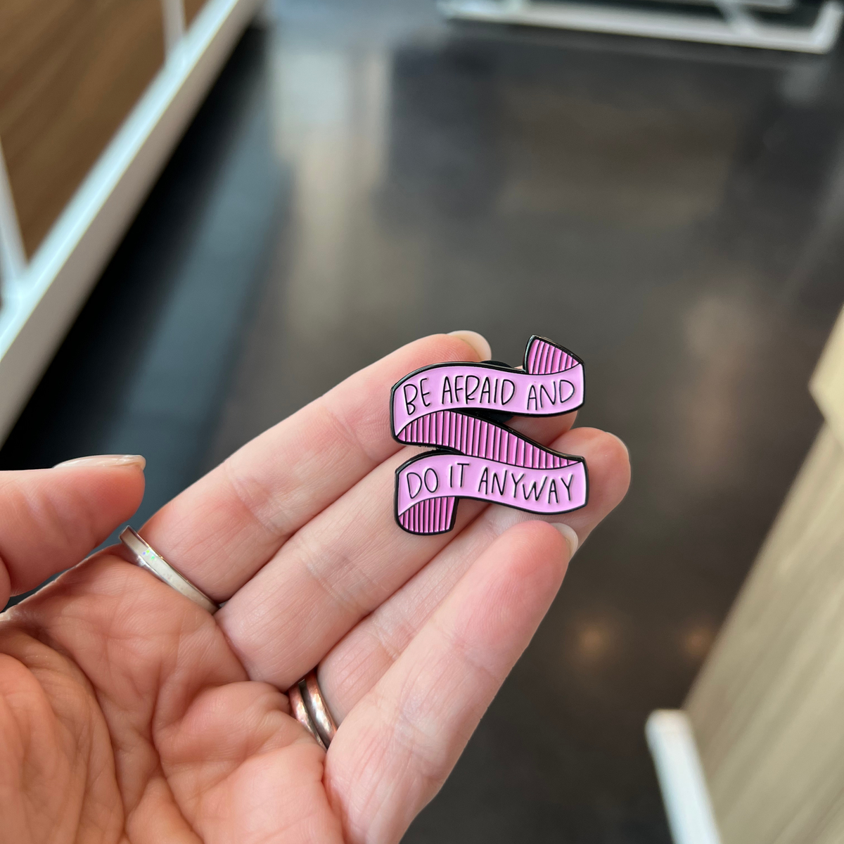 A hand holding a small enamel pin shaped like a ribbon with the text 'BE AFRAID AND DO IT ANYWAY' in black lettering on a pink striped background.