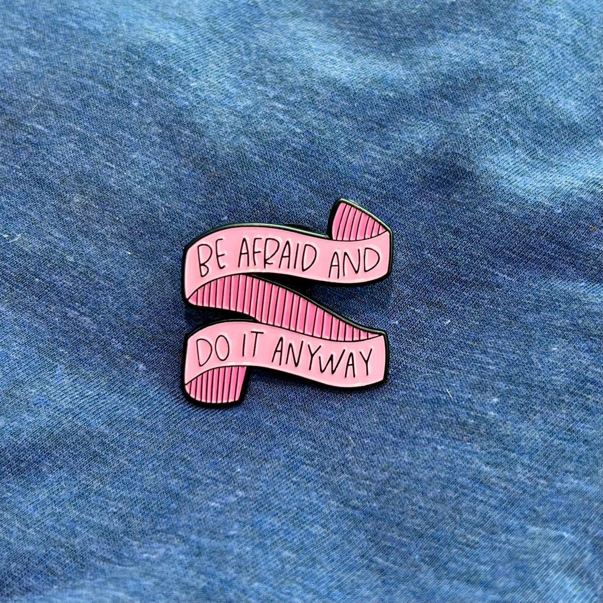 A pink enamel pin shaped like a ribbon banner with the text 'BE AFRAID AND DO IT ANYWAY' in black lettering, placed on a blue fabric background.