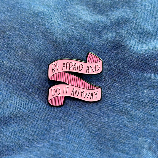 A pink enamel pin shaped like a ribbon banner with the text 'BE AFRAID AND DO IT ANYWAY' in black lettering, placed on a blue fabric background.