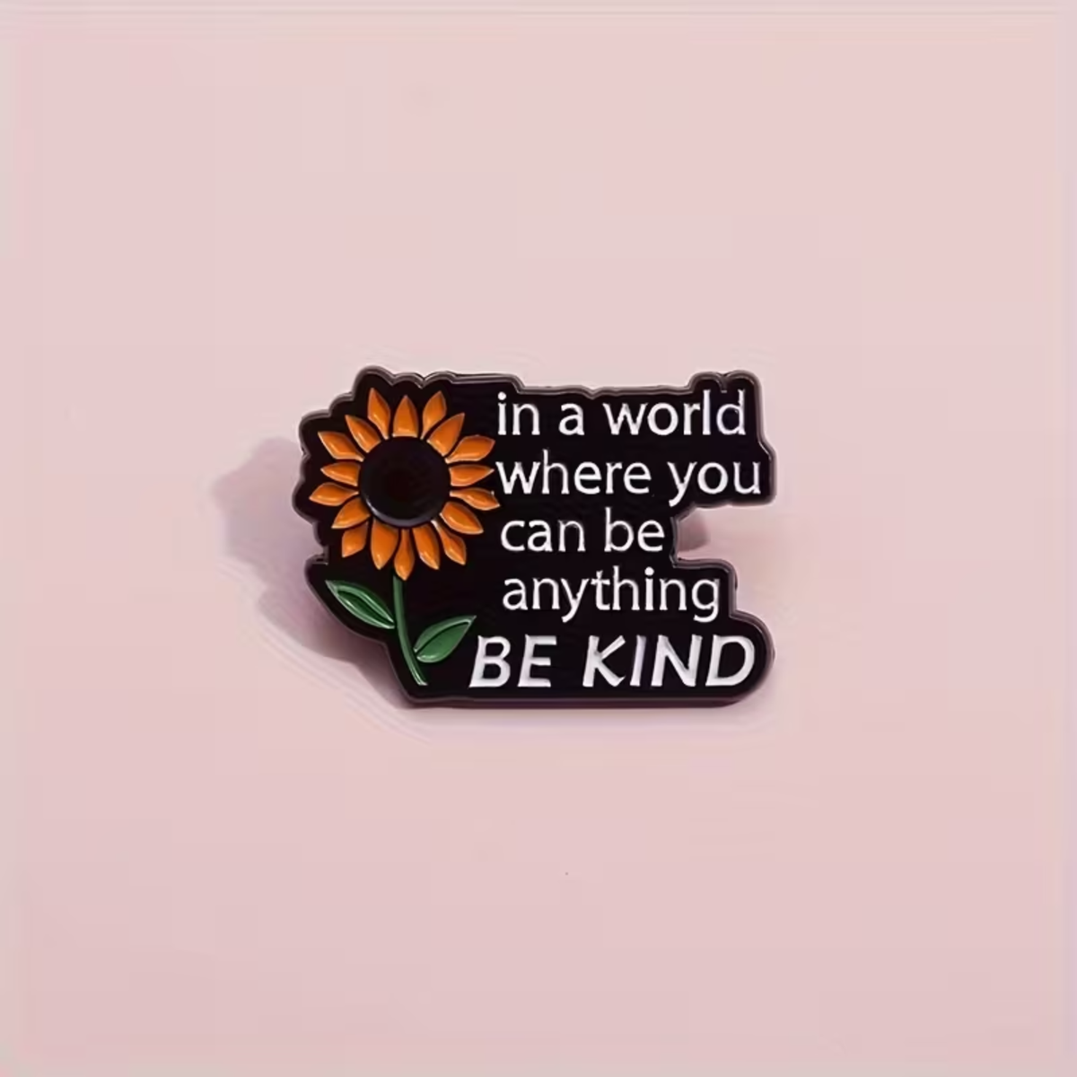 A small enamel pin shaped like a black speech bubble with a sunflower on the left and the phrase 'in a world where you can be anything BE KIND' in white text, shown on a pale pink background.