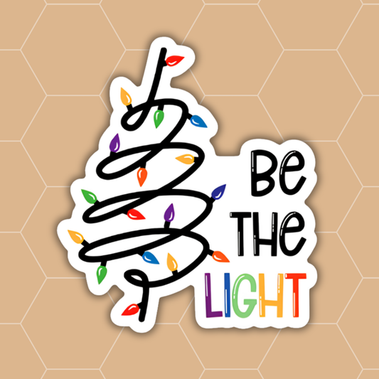 A die-cut sticker with a white border featuring a black spiral Christmas tree outline decorated with multicolored holiday lights and the words 'Be THE LIGHT' in black and rainbow lettering, shown on a tan hexagonal background.