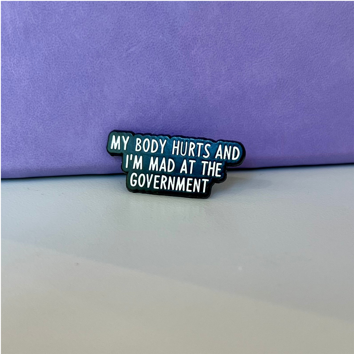 A small enamel pin with the text 'MY BODY HURTS AND I'M MAD AT THE GOVERNMENT' in white lettering on a black background with a purple outline, placed on a light surface against a purple backdrop.
