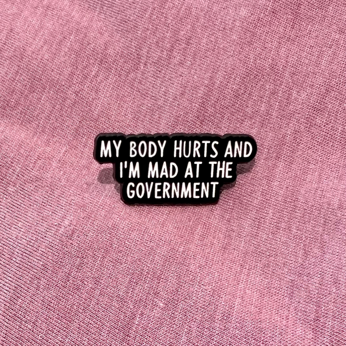 A black enamel pin with white text reading 'MY BODY HURTS AND I'M MAD AT THE GOVERNMENT' displayed on a textured pink fabric background.