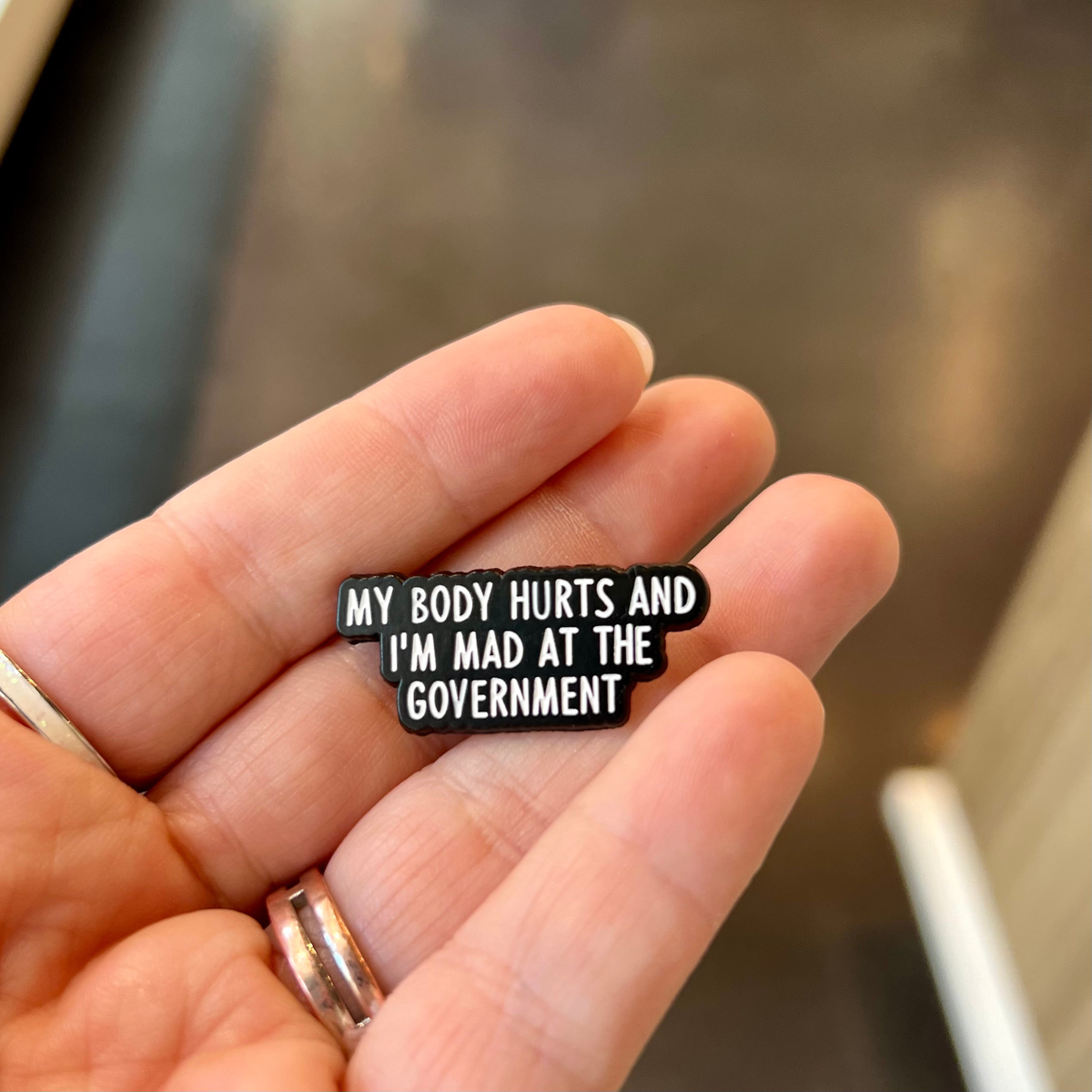 A close-up of a hand holding a small black enamel pin with white text that reads, 'MY BODY HURTS AND I'M MAD AT THE GOVERNMENT'.