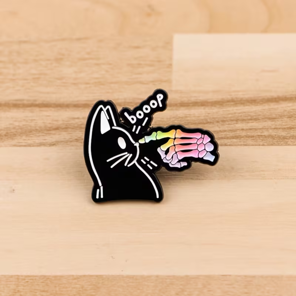 A black enamel pin featuring a cartoon cat with white outlines and a rainbow-colored skeleton hand touching its nose, with the word 'booop' above it, placed on a light wood surface.