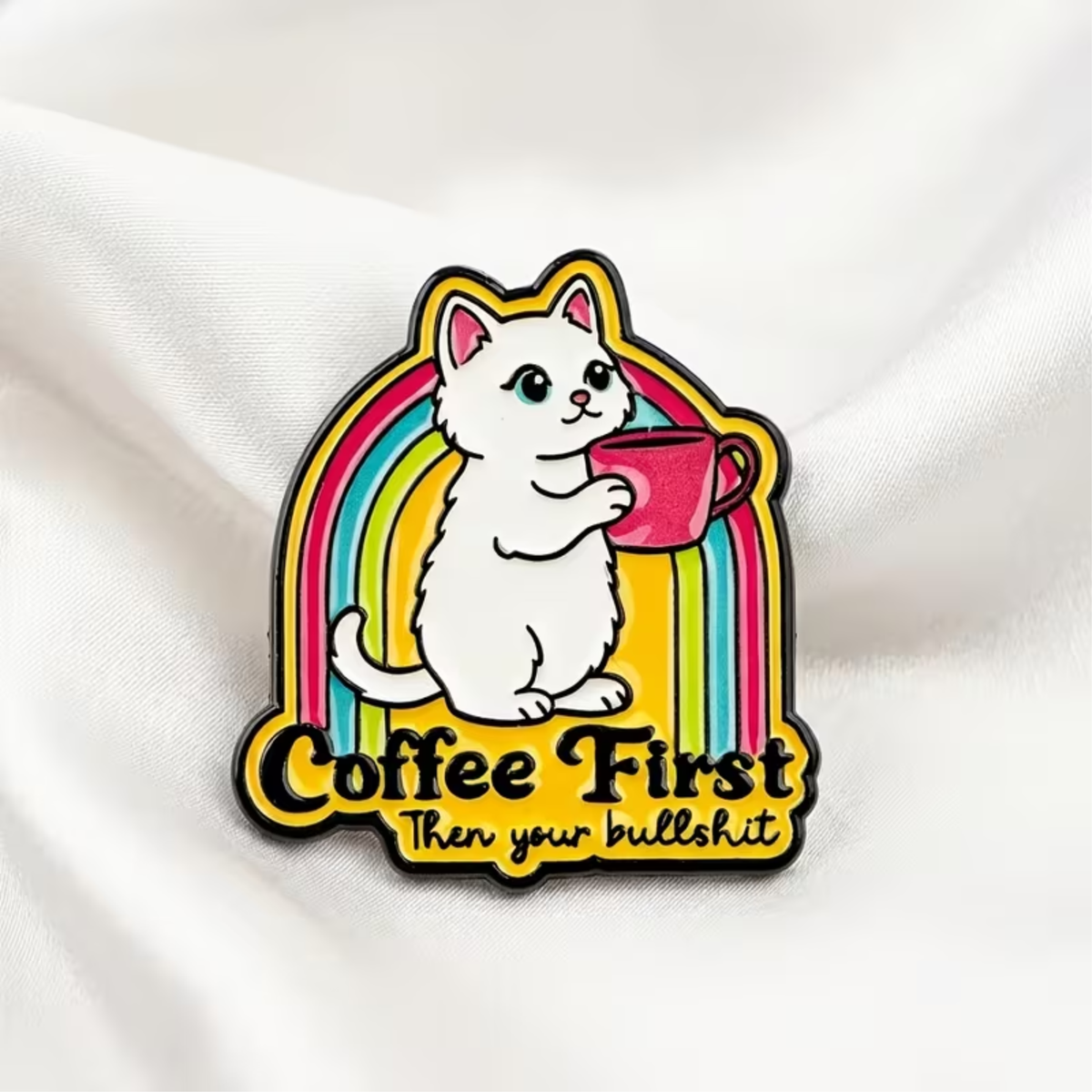 A colorful enamel pin featuring a cartoon white cat holding a pink coffee mug in front of a rainbow, with the text 'Coffee First Then your bullshit' on a white fabric background.