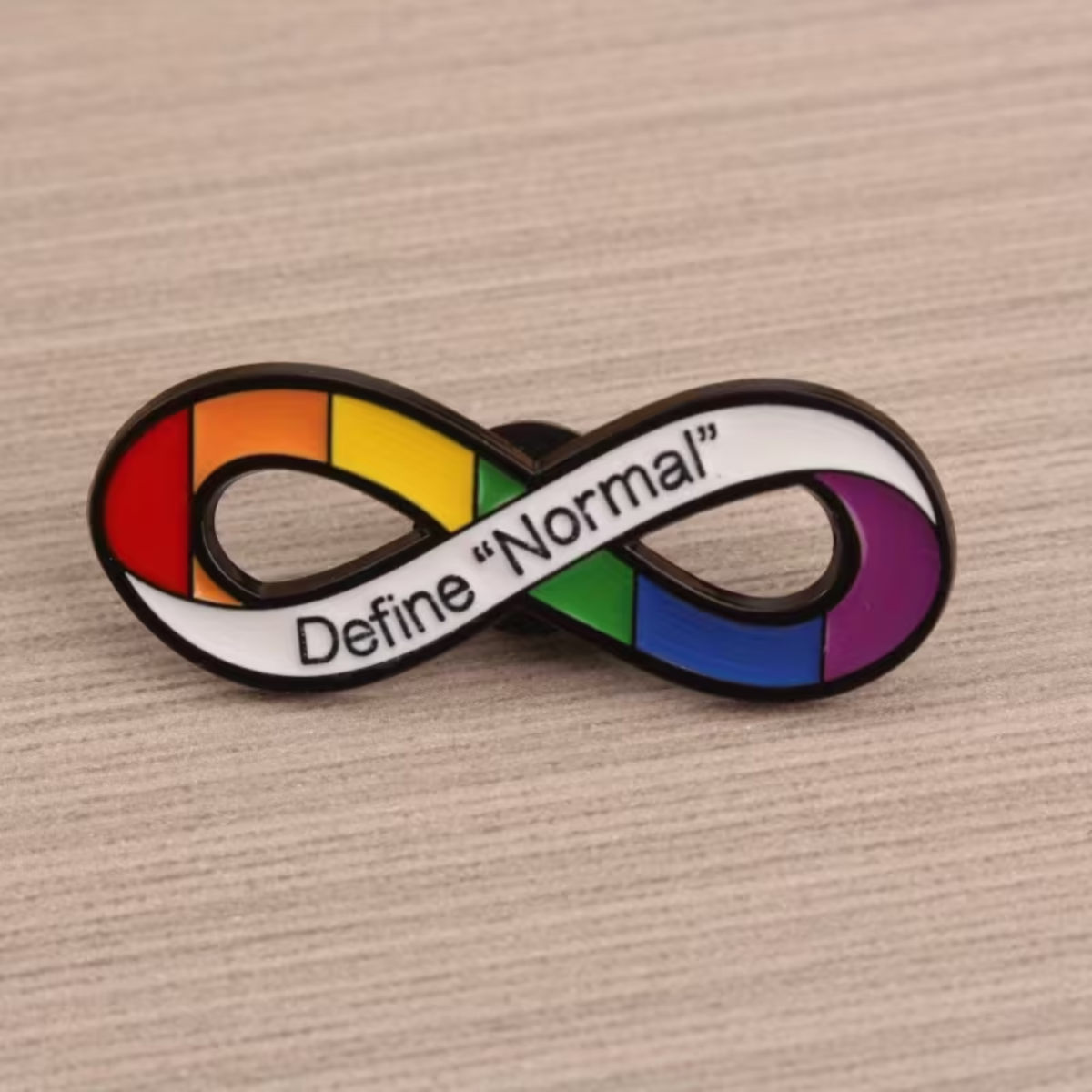 A rainbow pride infinity symbol lapel pin with the text 'Define "Normal"' on a white banner, displayed on a light wood surface.