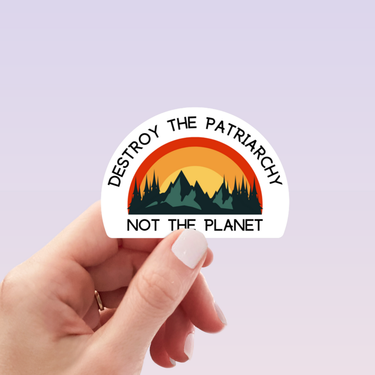 A hand with light pink manicured nails holds a die-cut sticker featuring a sunset landscape with mountains and the text 'DESTROY THE PATRIARCHY NOT THE PLANET' on a pale lavender background.