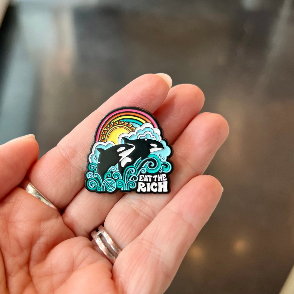 A hand holds an Eat the Rich Pin featuring two orcas leaping through stylized blue waves beneath a rainbow and sun.