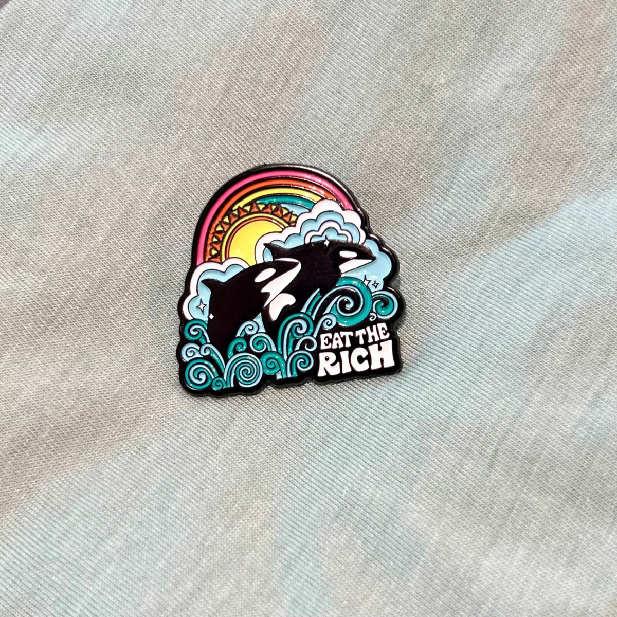 Eat the Rich Pin featuring two orcas jumping out of stylized waves in front of a sun and rainbow, with the text EAT THE RICH at the bottom of this activist accessory.