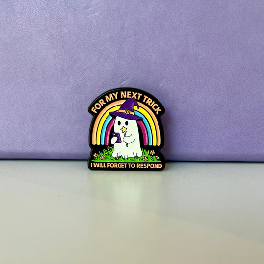 A small enamel pin featuring a white cat wearing a purple blindfold and rainbow hat, with the text 'FOR MY NEXT TRICK' above and 'I WILL FORGET TO RESPOND' below, displayed on a light surface against a lavender background.