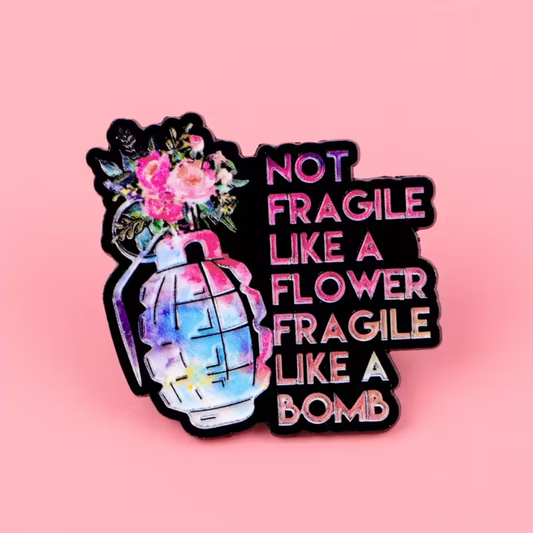 A novelty enamel pin shaped like a hand grenade with a watercolor-style floral burst and the text 'NOT FRAGILE LIKE A FLOWER FRAGILE LIKE A BOMB' in pink and white lettering on a black background, set against a solid pink backdrop.