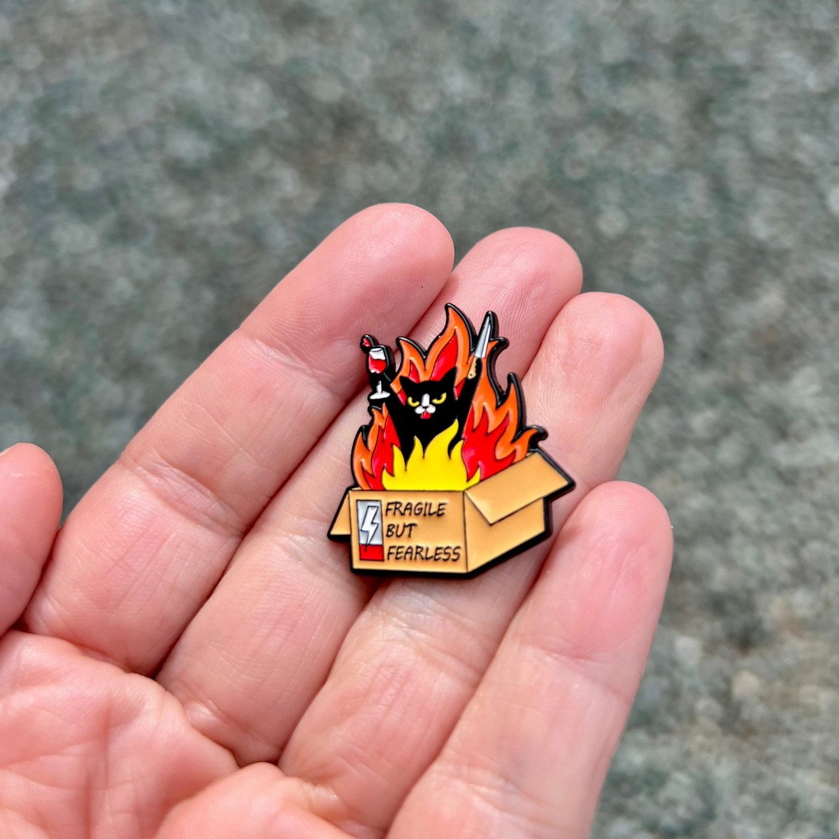 A Fragile But Fearless Pin rests in a palm, depicting a black cat holding a wine glass and knife inside a burning cardboard box labeled Fragile but fearless.