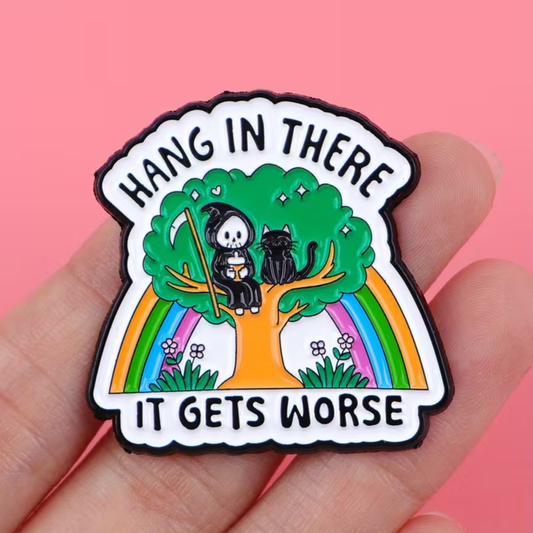 A hand holding a die-cut enamel pin that reads 'HANG IN THERE IT GETS WORSE' with a grim reaper, black cat, green tree, and rainbow on a pink background.