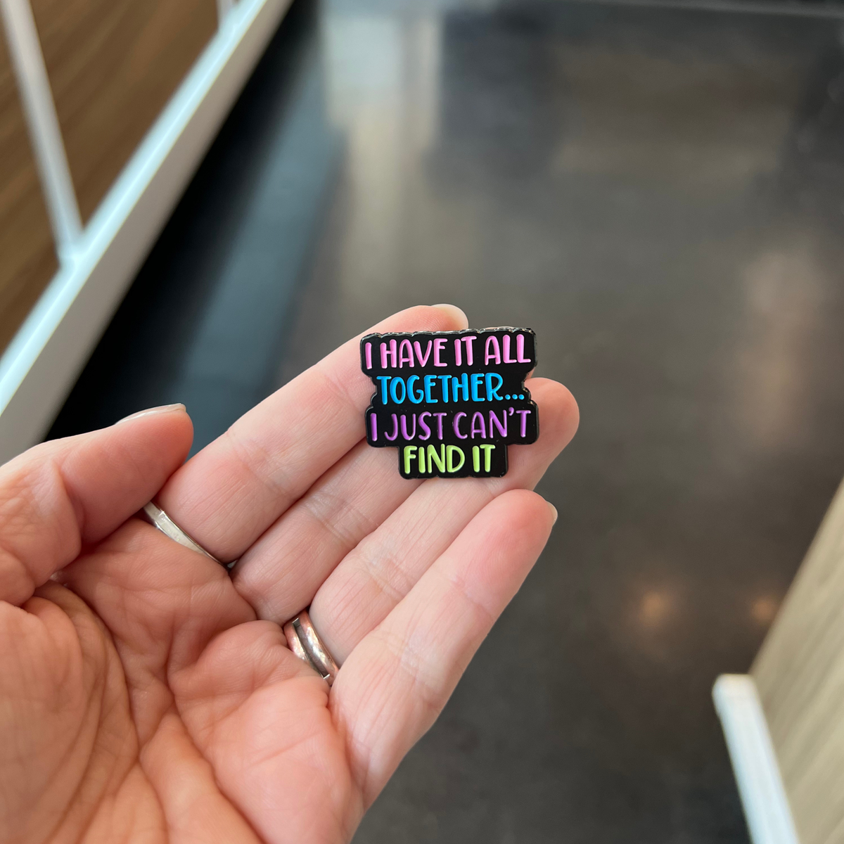A close-up of a hand holding a small enamel pin that reads 'I HAVE IT ALL TOGETHER... I JUST CAN'T FIND IT'