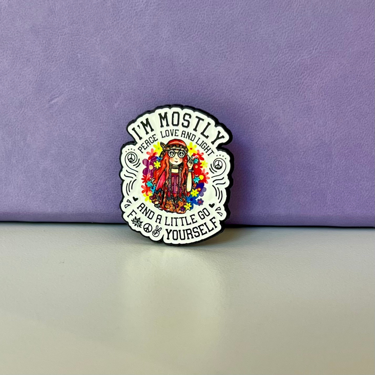 A die-cut enamel pin featuring a colorful illustration of a woman with glasses and the text 'I'M MOSTLY PEACE, LOVE AND LIGHT' and 'AND A LITTLE GO FUCK YOURSELF', displayed on a light gray surface against a textured purple background.