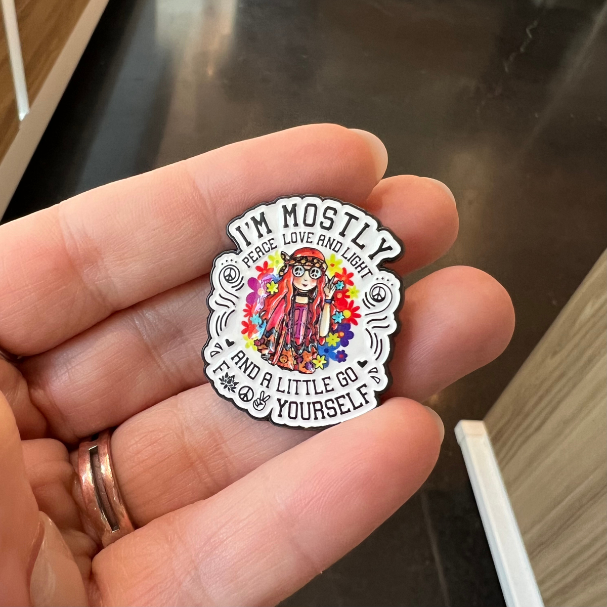 A hand holding a small enamel pin with a colorful illustration of a woman surrounded by flowers and the text 'I'M MOSTLY PEACE LOVE AND LIGHT AND A LITTLE GO FUCK YOURSELF'.