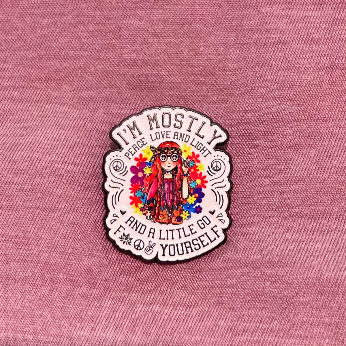 A die-cut enamel pin featuring a colorful cartoon girl with glasses surrounded by flowers, with the text 'I'M MOSTLY PEACE LOVE AND LIGHT AND A LITTLE GO FUCK YOURSELF' on a pink textured fabric background.