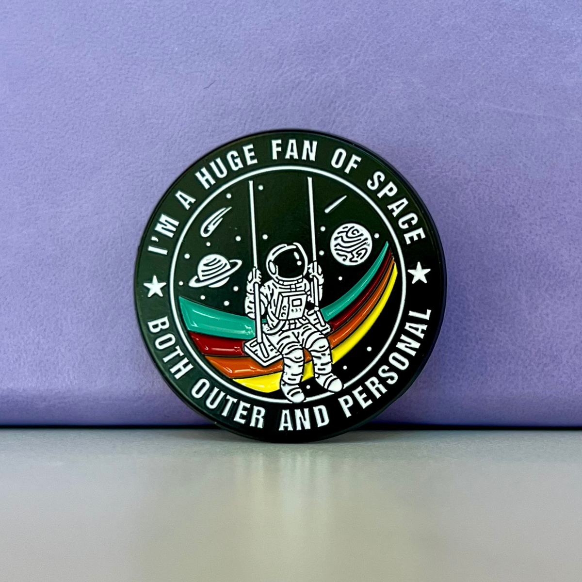A round enamel pin featuring an astronaut on a swing with planets and stars, bordered by the text 'I'M A HUGE FAN OF SPACE BOTH OUTER AND PERSONAL'. The pin is displayed on a light gray surface against a textured purple background.