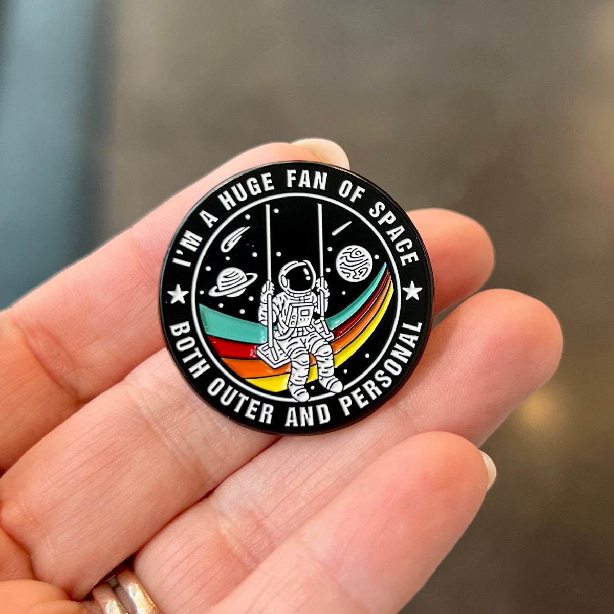 A circular enamel pin featuring a white astronaut graphic on a black background with colorful rainbow stripes and space-themed illustrations. The pin is held in a person's hand, with the text 'I'M A HUGE FAN OF SPACE BOTH OUTER AND PERSONAL' printed around the edge.