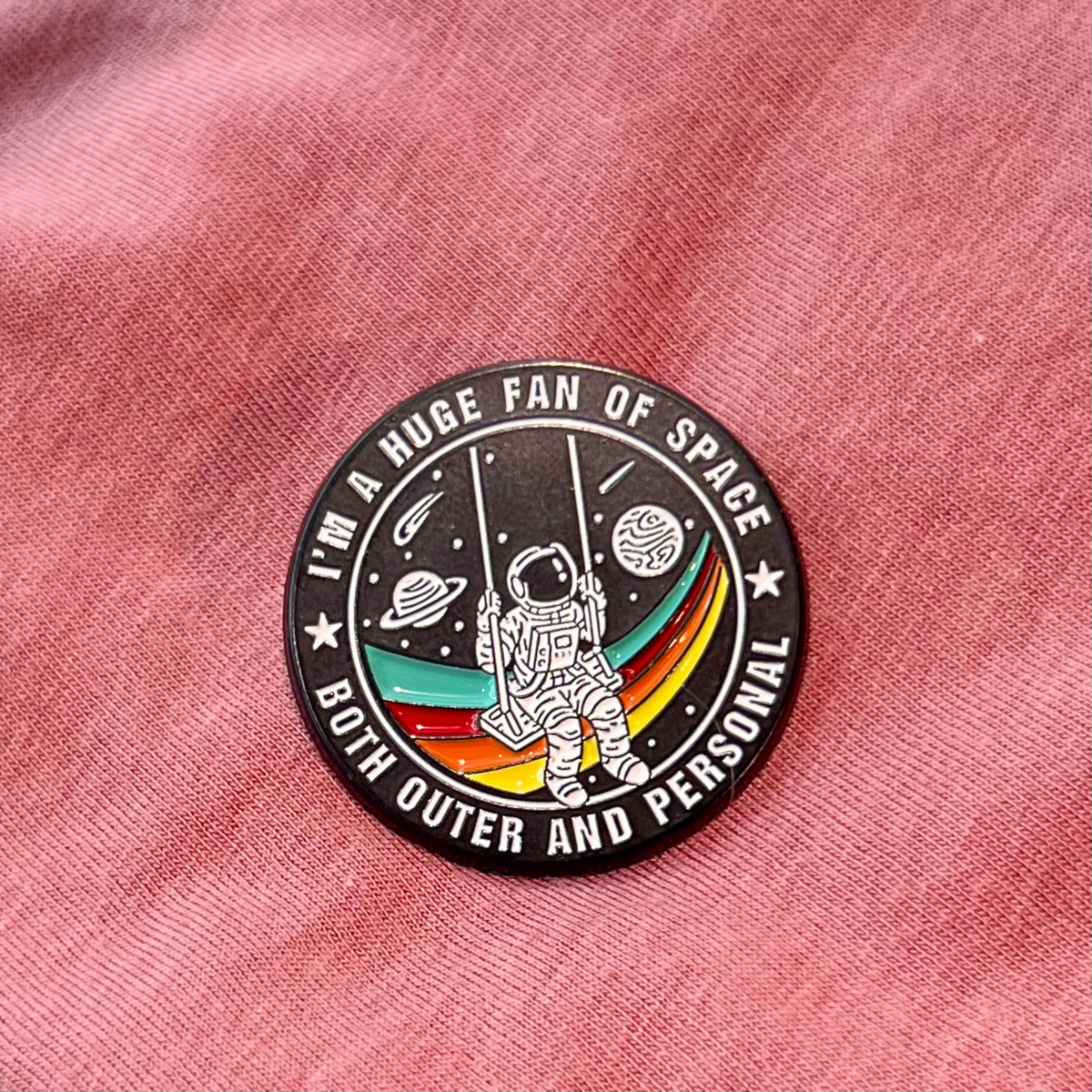 A circular enamel pin featuring an astronaut and planets on a black background with the text 'I'M A HUGE FAN OF SPACE BOTH OUTER AND PERSONAL', placed on a pink textured fabric background.