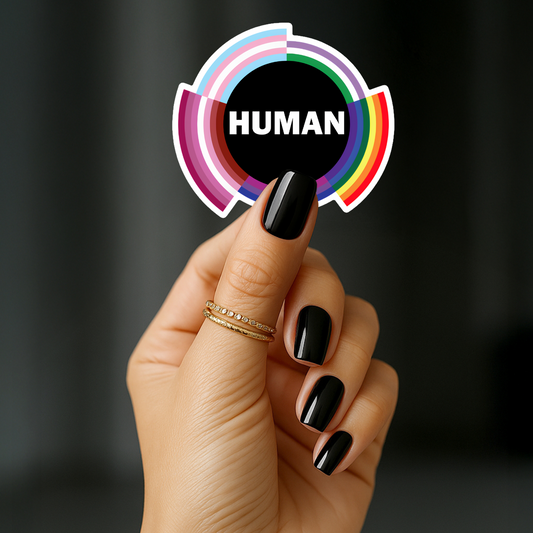 A hand adorned with black nail polish and gold rings holds the HUMAN Sticker by RainBee Designs LLC—a circular sticker featuring rainbow borders and the word "HUMAN" in bold white letters at the center.