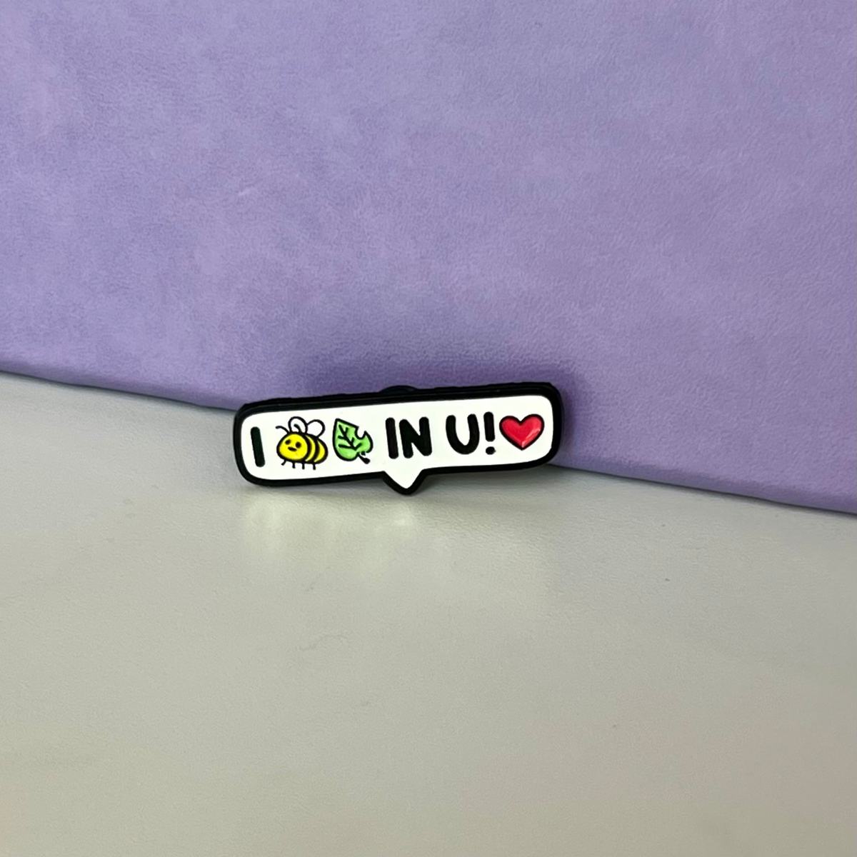 A small enamel pin shaped like a speech bubble with the text 'I (bee icon and leaf icon - read as BELIEVE) IN U!'. It features a white background, a black outline, a yellow cartoon bee, a green leaf, and a red heart.