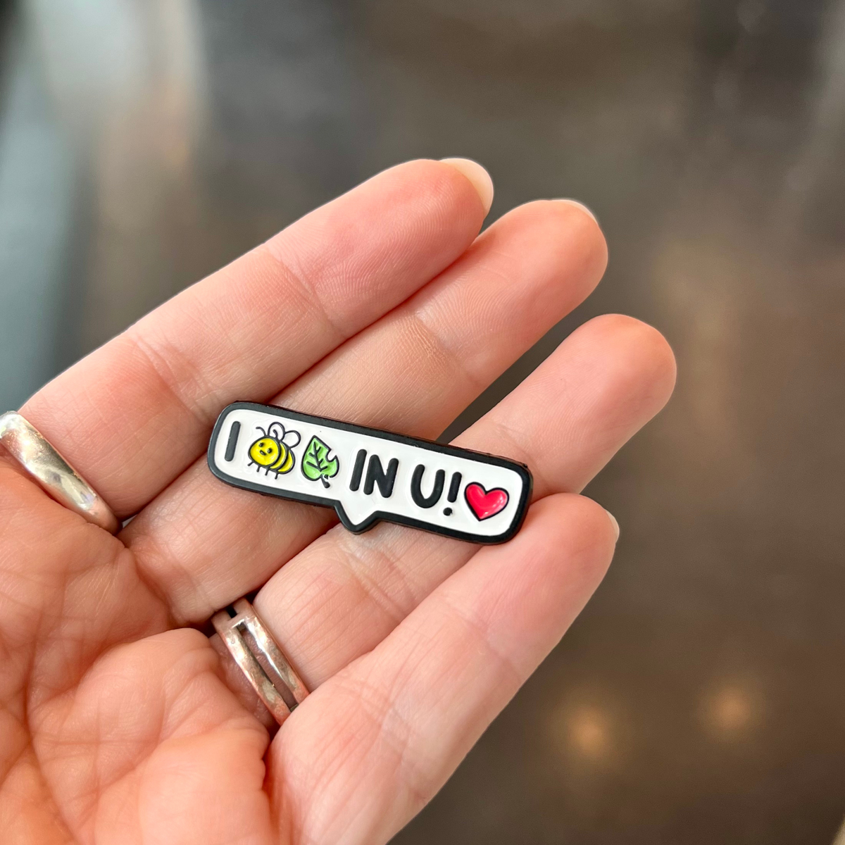 A small enamel pin shaped like a speech bubble is held on a person's fingers. The pin has a white background with a black outline and the text 'I (bee icon and leaf icon - read as BELIEVE) IN U!' accompanied by a yellow bee, green leaves, and a red heart.